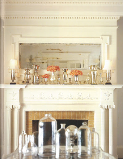 On the shelf of a mirrored mantelpiece, reflective mercury-glass lamps, vases, and other vessels provide a warm, luminous setting for the roses that bring the only touch of intense color.