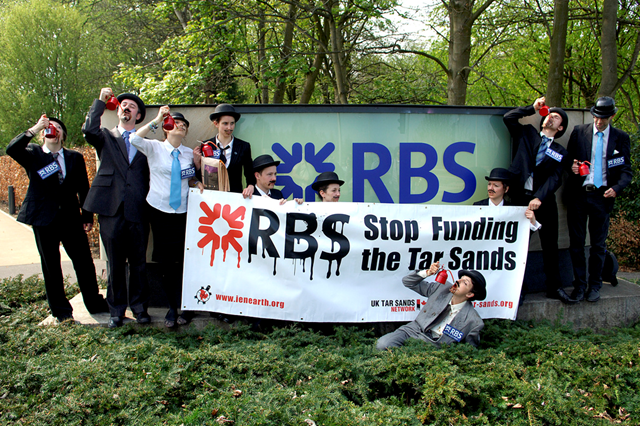 Environmental campaigners dressed as bankers protest outside the RBS Headquarters at Gogarburn in Edinburgh ahead of the bank's AGM, 19 April 2011. Human rights and environmental campaigners dressed as oil-addicted bankers drinking from oil cans, demanded an end to the bank's support for the fossil fuel industry. They were joined by several Canadian First Nations representatives who arrived to personally urge for RBS to cease financing tar sands companies extracting oil on their lands. Photo: Richard Scott / MAVERICK