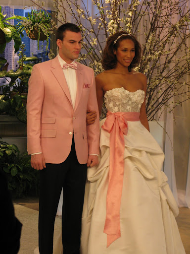 A Monique Lhuillier gown with meringue texture is shown here while the groom is showing off a pink jacket and black pants from Kleinfeld.