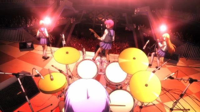 An all-female band plays on a red-lit stage before a huge crowd, scene from just above the drummer's head looking out