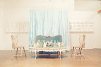 An intimate lounge area in front of a soft blue fabric backdrop boasts an eclectic mix of furniture styles. The art piece in the background is An intimate lounge area in front of a soft blue fabric backdrop boasts an eclectic mix of furniture styles. The art piece in the background is