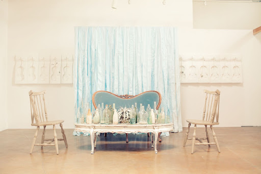 An intimate lounge area in front of a soft blue fabric backdrop boasts an eclectic mix of furniture styles. The art piece in the background is An intimate lounge area in front of a soft blue fabric backdrop boasts an eclectic mix of furniture styles. The art piece in the background is