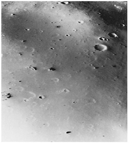 View of the Martian moon Deimos taken on October 15, 1977 when Viking Orbiter 2 passed within 50 kilometers (30 miles) of the satellite. The picture covers an area 1.2 x 1.5 kilometers (0.74 x 0.93 mile) and shows features as small as 3 meters (10 feet). Deimos is saturated with craters, but a layer of dust appears to cover craters smaller than 50 meters (165 feet) in diameter, making Deimos look smoother than the other Martian moon, Phobos. Boulders as large as houses (10 to 30 meters; 33 to 100 feet) across are strewn about the face. It is suggested that these objects may be blocks ejected from nearby craters. The spacecraft would have been clearly visible to an observer standing on the surface of Deimos.