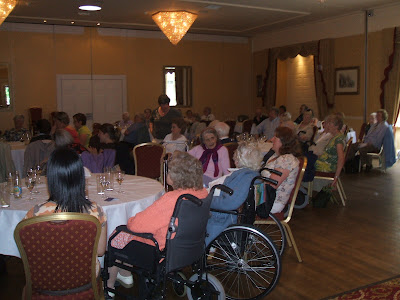 shot of many people, some old, some in wheelchairs, at hotel tables all looking at a stage