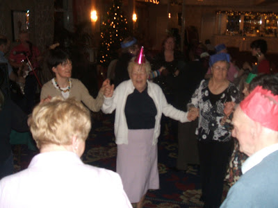 A group of old people dressed in party hats in a circle up for a dance in a big room. Loads of people up dancing