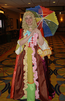 Young woman in a variant Sixth Doctor costume that has been re-imagined as an 18th century ballgown. She is carrying a brightly colored parasol and has a wig of blonde sausage curls.