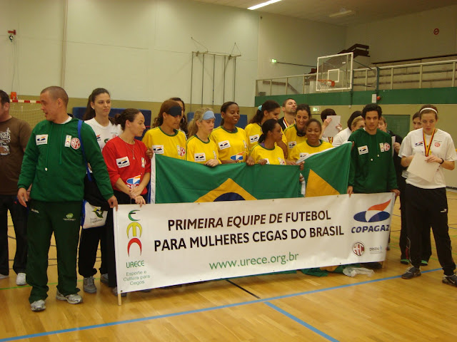 Na cerimônia de premiação, a equipe está dentro de quadra,segurando um cartaz onde se lê "Primeira Equipe de Futebol para mulheres  cegas do Brasil"