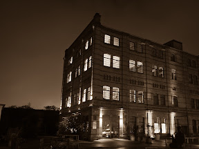 [ASSADE EINES BACKSTEINFABRIKGEBÄUDES IN BERLIN KREUZBERG. NACHTS, DIE RÄUME IM DEN FABRIKETAGEN SIND BLAU ERLEUCHTET. IM HINTERGRUND LEUCHTET DIE STADT. DAS BILD IST IN SEPIA GEHALTEN.]