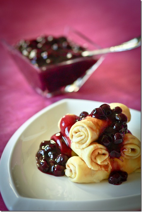 Farmer’s cheese filled crepes rolled and stacked on a plate drizzle with a homemade blueberry sauce. 