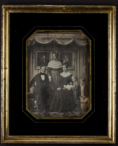 Family of three adults and three children in front of paintings of the man's parents