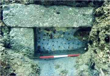 A sluice gate for a channel controlling the flow of seawater into a fish tank on Ventotene Island, south of Rome, with a modern-day red and white measuring rod. The gate is made of limestone with holes to allow flow during high tide, and it can slide vertically into the stone blocks to the left and right. When it was operating 19 centuries ago, low tide occurred at the level of the base of the sliding block. Today the entire gate is below sea level.
