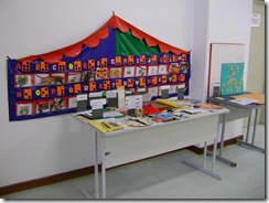 Material tatil para ensino de Braille disposto numa tenda colorida presa na parede. Mesa com recursos para ensino de pessoas com deficiência visual