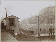 Mannen, vermoedelijk werknemers, en twee katten bij een aanlegsteiger en een wachthuisje langs een gracht