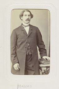 Studioportret van een staande man met snor bij een tafel, hij draagt een lange jas