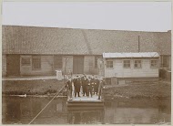 Mannen, vermoedelijk werknemers, staand op een aanlegsteiger bij een fabriekshal