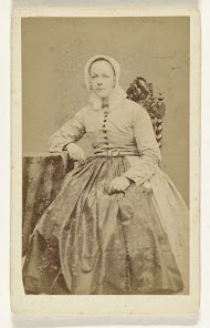 Studioportret van een vrouw in bolle rok met kap op het hoofd, zittend aan tafel